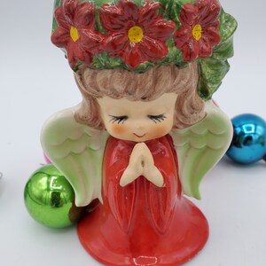 May include: A ceramic angel figurine with red poinsettia flowers on its head, wearing a red dress and praying with its hands clasped. The angel has green wings and a sweet expression on its face.
