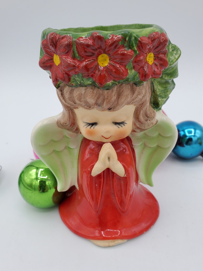 May include: A ceramic angel figurine with red poinsettia flowers on its head, wearing a red dress and praying with its hands clasped. The angel has green wings and a sweet expression on its face.
