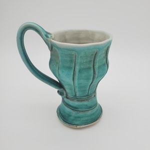 May include: A handmade turquoise ceramic mug with a unique, fluted design. The mug features a sturdy handle and a wide, off-white interior. The exterior has vertical grooves and a textured finish, adding to its artisanal appeal. A decorative piece for the kitchen.
