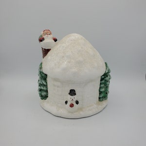 May include: White ceramic Christmas figurine of a snow-covered house with a snowman in front and Santa Claus peeking over the roof.