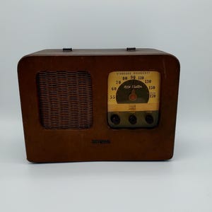 Puede incluir: Radio RCA Victor vintage en una caja de madera marrón. La radio presenta una cubierta de altavoz tejida, un dial de sintonización con números y el texto "STANDARD BROADCAST". Tiene tres perillas y el logotipo de RCA Victor.