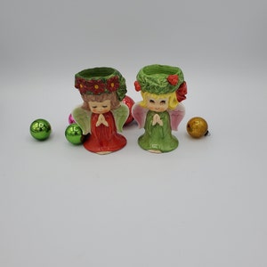 May include: Two ceramic angel candle holders with green and red floral wreaths on their heads. The angels are wearing red and green dresses and have pink wings. They are holding their hands together in a prayer position. There are green and gold ornaments in the background.