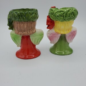 May include: Two ceramic angel figurines with green wreaths on their heads. One angel is wearing a red dress and the other is wearing a green dress. Both angels have pink wings.
