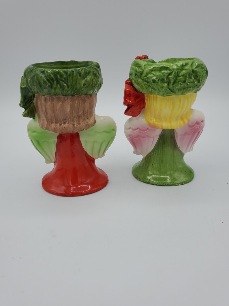 May include: Two ceramic angel figurines with green wreaths on their heads. One angel is wearing a red dress and the other is wearing a green dress. Both angels have pink wings.