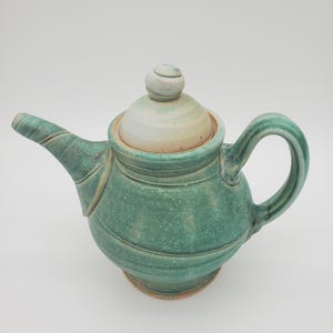May include: A handmade ceramic teapot in shades of green and off-white. The teapot features a rounded body with horizontal ridges, a curved spout, and a loop handle. The lid has a small, rounded knob. The teapot is a unique piece of pottery.