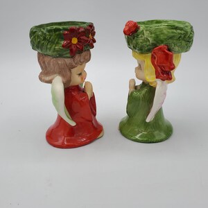 May include: Two ceramic angel candle holders with green wreaths on their heads. One angel is wearing a red dress and the other is wearing a green dress.