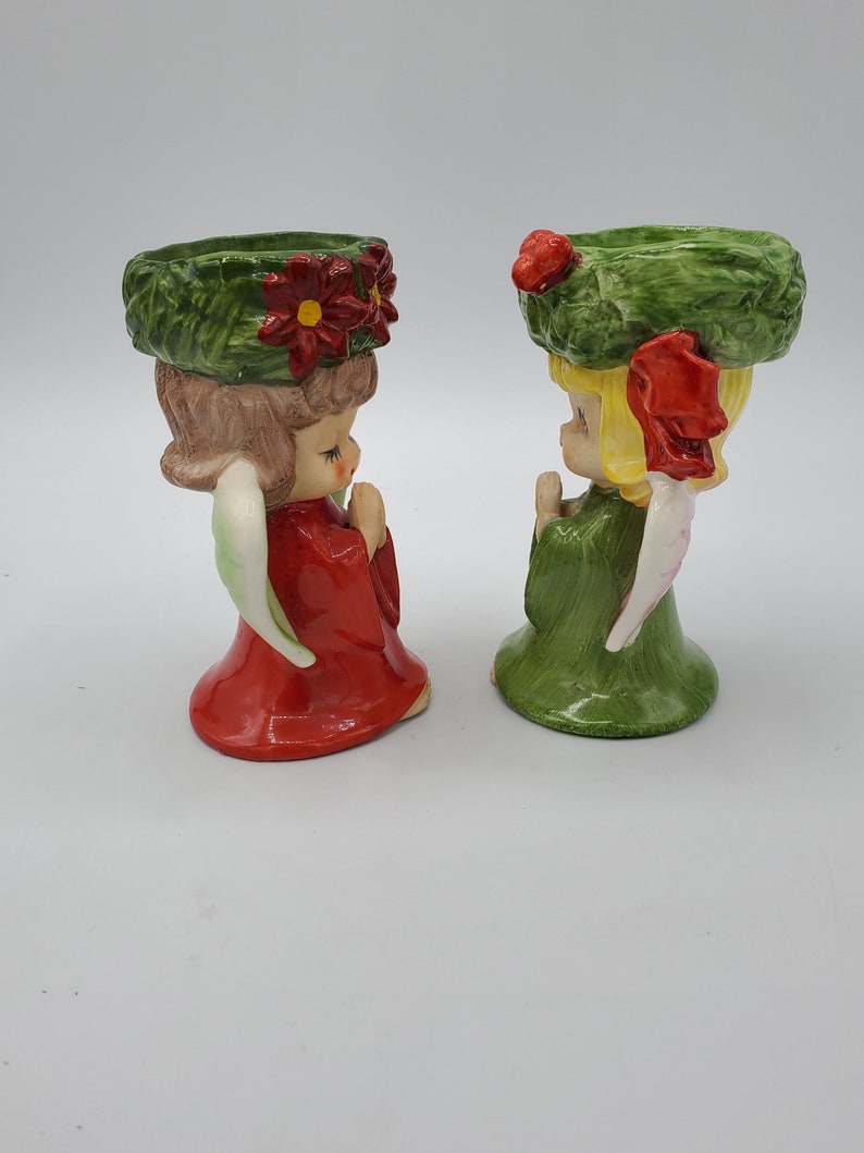 May include: Two ceramic angel candle holders with green wreaths on their heads. One angel is wearing a red dress and the other is wearing a green dress.