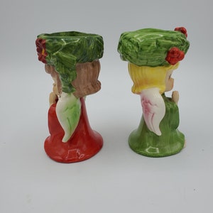 May include: Two ceramic angel candle holders with green wreaths on their heads. One angel is wearing a red dress and the other is wearing a green dress.