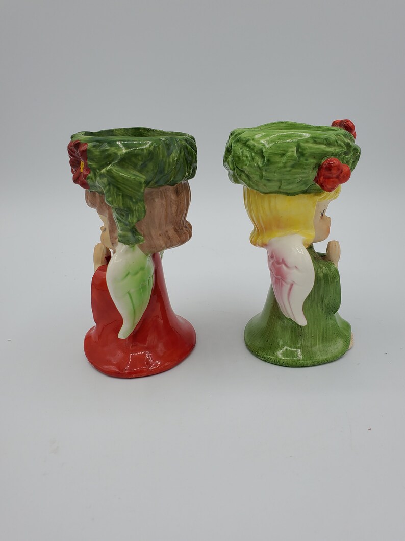 May include: Two ceramic angel candle holders with green wreaths on their heads. One angel is wearing a red dress and the other is wearing a green dress.