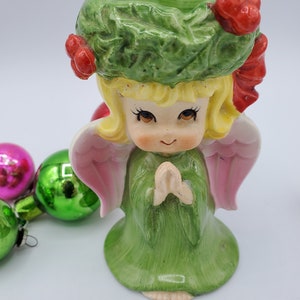 May include: A green and pink ceramic angel figurine with a wreath on its head and its hands clasped in prayer. The angel is wearing a green dress and has a pink halo.