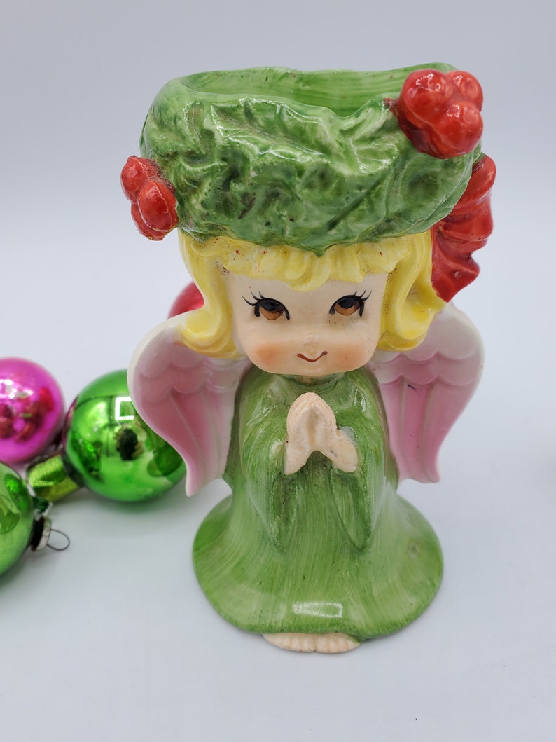 May include: A green and pink ceramic angel figurine with a wreath on its head and its hands clasped in prayer. The angel is wearing a green dress and has a pink halo.