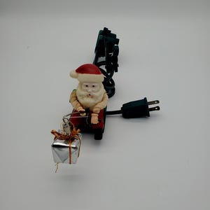 May include: A Santa Claus figurine wearing a red hat and suit, holding a fishing rod with a silver gift box attached to the line. The figure is connected to a black electrical cord with a plug, featuring a dark green base and light accents.