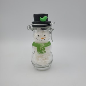 May include: A snowman-shaped glass jar with a black top hat and a green bird decoration. The jar has a green scarf and a face made of dots and a small orange carrot nose. The jar is filled with white tissue paper.