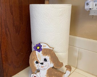 May include: A white paper towel roll with a wooden topper. A decorative, hand-painted wooden puppy dog is attached to the roll. The puppy has brown and white fur, a purple flower, and is standing on green grass.