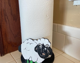 May include: A black and white sheep paper towel holder with a green base. The holder is made of wood and has a black knob on top. The sheep is painted with a black face and white fur. The holder is sitting on a white tiled surface.