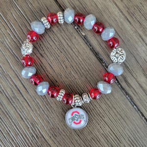 May include: A beaded bracelet featuring red and gray beads with silver accents. The bracelet includes a charm with the Ohio State logo. The beads are strung on a stretchy cord.