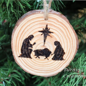 May include: A round, wooden Christmas ornament with a rustic aesthetic. The ornament features a pyrography design of the Nativity scene, including Mary, Joseph, the baby Jesus, and the star of Bethlehem. The text "Burning Creations" is at the bottom.