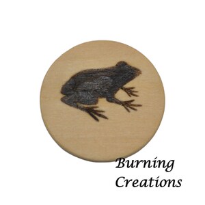 May include: A round wooden disc with a woodburned image of a frog. The disc is light brown with a dark brown frog silhouette. The text "Burning Creations" is printed below the disc.