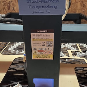 May include: A black laser engraving machine with a blue top, featuring the text "Mad-Mitten Engraving Jackson, Mi." The machine has a "LONGER" label with technical specifications and safety warnings. The machine is designed for engraving.