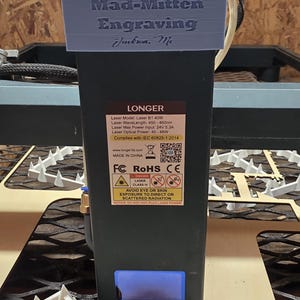 May include: A black laser engraving machine with a blue top, featuring the text "Mad-Mitten Engraving Jackson, Mi." and "LONGER." The machine has safety labels and technical specifications, including laser wavelength and power.
