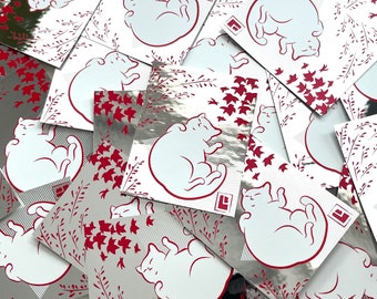 May include: A stack of silver holographic stickers featuring a white cat curled up with red floral designs and red leaves.
