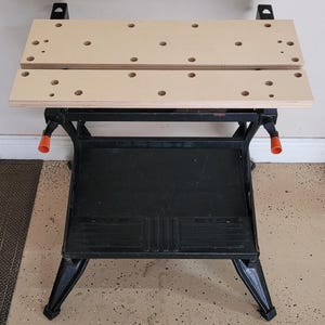 May include: A black metal workbench with two wooden work surfaces. The work surfaces have multiple holes drilled into them. The workbench has a lower shelf and adjustable legs.
