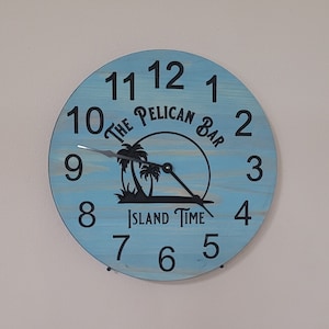 May include: A blue wooden clock with black numbers and text that reads "The Pelican Bar Island Time". The clock has a silhouette of palm trees and a sun.
