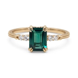 Peut inclure: Une bague en or avec une pierre précieuse vert foncé taille émeraude fixée par quatre griffes. L'anneau est orné de deux petites pierres rondes transparentes de chaque côté et présente un détail de bordure texturé de style milgrain.