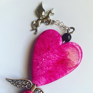 May include: A pink heart-shaped charm with a silver winged unicorn charm attached to it. The heart charm has a white and pink marbled design. The unicorn charm is attached to the heart charm with a silver chain.