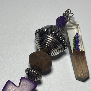 May include: A purple cross charm with a silver bead, a brown wooden bead, a silver bead with a textured surface, a purple skull bead, and a clear resin charm with a purple flower and a wood accent.
