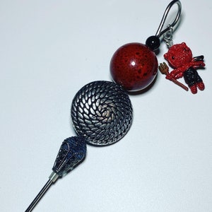 May include: A silver metal button with a textured design, a red ceramic bead, a small red devil figurine, and a black bead on a silver metal hook.
