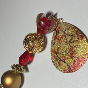 May include: A gold and red beaded ornament with a heart-shaped charm, a round bead with a filigree design, and a red crystal bead. The ornament also features a gold leaf-shaped charm with a red and gold design.