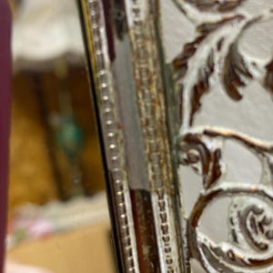 May include: A silver-toned metal mirror frame featuring an intricate openwork design with scrolling leaf patterns. The frame has a metallic finish with visible signs of aging, including dark patina and wear along the textured edges.