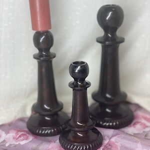 May include: Three dark brown wooden candle holders of varying heights. One holds a pink candle. The candle holders have a turned design with a round base and a decorative top. The candle holders are displayed on a floral patterned fabric.