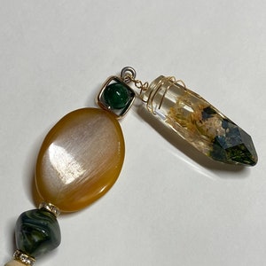 May include: A close-up of a necklace with a variety of beads and a clear, faceted pendant. The necklace features a large, oval, amber-colored bead, a square green bead, and a clear pendant with dried flowers inside. The necklace is on a white background.