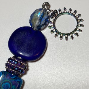 Puede incluir: Un collar de cuentas azul y morado con un dije plateado en forma de sol con diamantes de imitación. El collar tiene una variedad de cuentas, incluyendo una cuenta redonda azul grande, una cuenta redonda azul más pequeña y una sección de cuentas con cuentas moradas y plateadas.