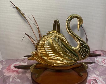 May include: A gold swan sculpture with a green and gold patterned design. The swan is perched on a wooden base with three legs. The swan has a long neck and a small head with a blue beak. There are three thin copper-coloured reeds behind the swan.