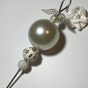 May include: White beaded ornament with a pearl, a white ceramic angel with wings, and a silver hook. The ornament is designed to hang on a Christmas tree.