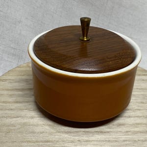 May include: A round ceramic container with a smooth, solid mustard-yellow finish and a white interior rim. The container features a circular dark brown wooden lid topped with a small, centered, metallic gold-toned conical handle.