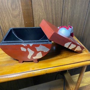 May include: A red and black wooden box with a lid, featuring silver and gold accents. The box has a unique shape and is open, revealing a black interior. The lid has a decorative element and a red tassel.