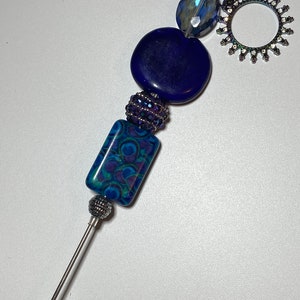 May include: A blue and green patterned glass bead, a dark blue glass bead, a sparkly purple bead, and a clear glass bead are strung on a silver metal rod. The rod is topped with a silver metal sun-shaped charm with a circle in the center.