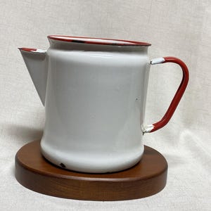 May include: White enamelware pitcher with a red rim and handle. The pitcher sits on a round, wooden base. The spout is triangular, and the handle is a curved D-shape. The pitcher has a vintage aesthetic.