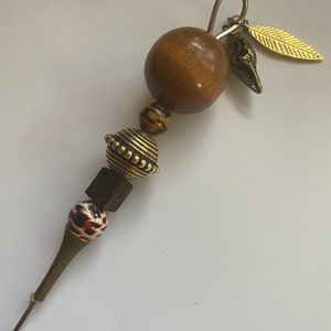 May include: A brown wooden bead with gold and black beads, a brown cube, and a brown and white patterned bead. The beads are strung on a metal wire with a gold leaf and a shell charm.