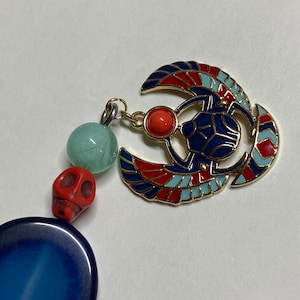 May include: A gold-colored pendant featuring a blue scarab beetle with red and blue wings. The pendant is attached to a chain with green and red beads.