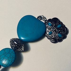 May include: A turquoise heart-shaped pendant with a silver filigree charm and turquoise accents. The pendant is part of a turquoise and silver necklace.