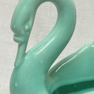 Puede incluir: Una maceta de cerámica en forma de cisne de color verde menta claro brillante, con un cuello largo y curvado y un acabado liso y reflectante. El cisne está esculpido con un centro hueco, mostrando una forma continua y fluida de tono suave.