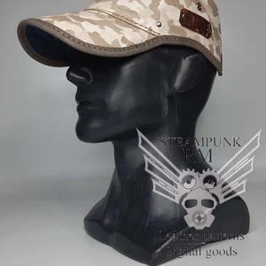 Puede incluir: Una gorra de béisbol de cuero beige con estampado de camuflaje. La gorra tiene una visera de cuero marrón y una correa de cuero marrón con una hebilla de metal. La gorra tiene un pequeño parche de cuero en la parte superior con el texto "STEAMPUNK RM" en negro.