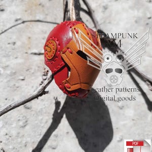 Könnte beinhalten: Ein rotes und braunes Leder-Steampunk-Helm-Ornament mit einem gestickten Design. Das Ornament hängt an einem Ast. Der Text "STEAMPUNK RM" und "Leather patterns Digital goods" ist auf dem Bild zu sehen. Ein PDF-Download-Symbol ist ebenfalls sichtbar.