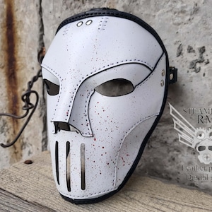 May include: White leather mask with a black strap and red paint splatters. The mask has a large, open mouth with a metal grill and a small, pointed nose.