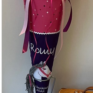 Könnte beinhalten: Ein rosa-violetter Schultüte mit weißen Punkten und dem Namen "Romy" bestickt. Die Tüte ist mit rosa und violetten Bändern verziert und hat ein kleines Stoffpferdchen daran befestigt. Die Tüte steht auf einem Metallständer und im Hintergrund ist eine braune Lederschultasche mit einem blauen Stern-Aufkleber zu sehen, auf dem "Stars ByLeAn" steht.